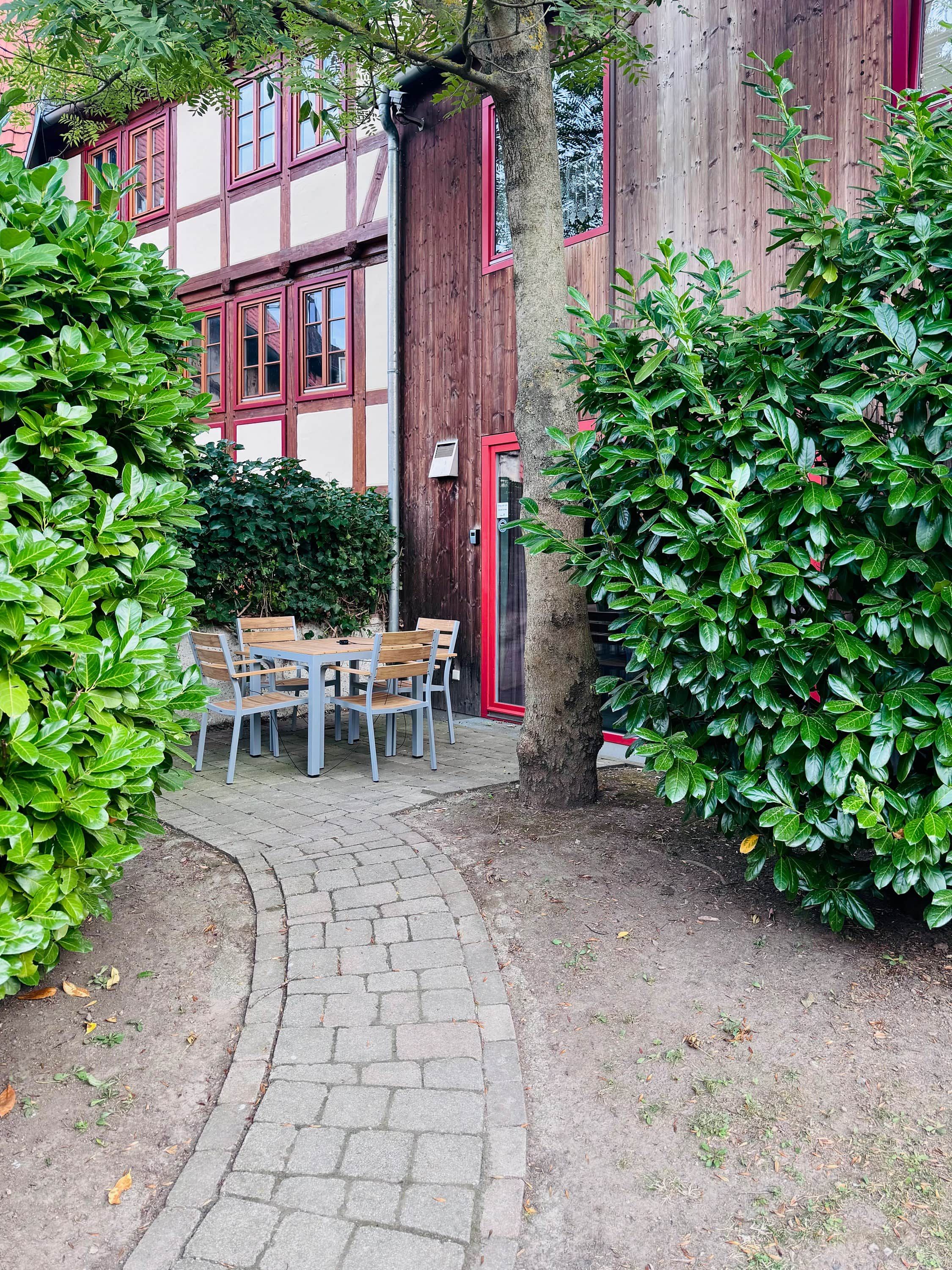 Une cour avec une table et des chaises en bois, entourée de buissons verdoyants et bordée d’un chemin pavé.