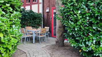 Une cour avec une table et des chaises en bois, entourée de buissons verdoyants et bordée d’un chemin pavé.