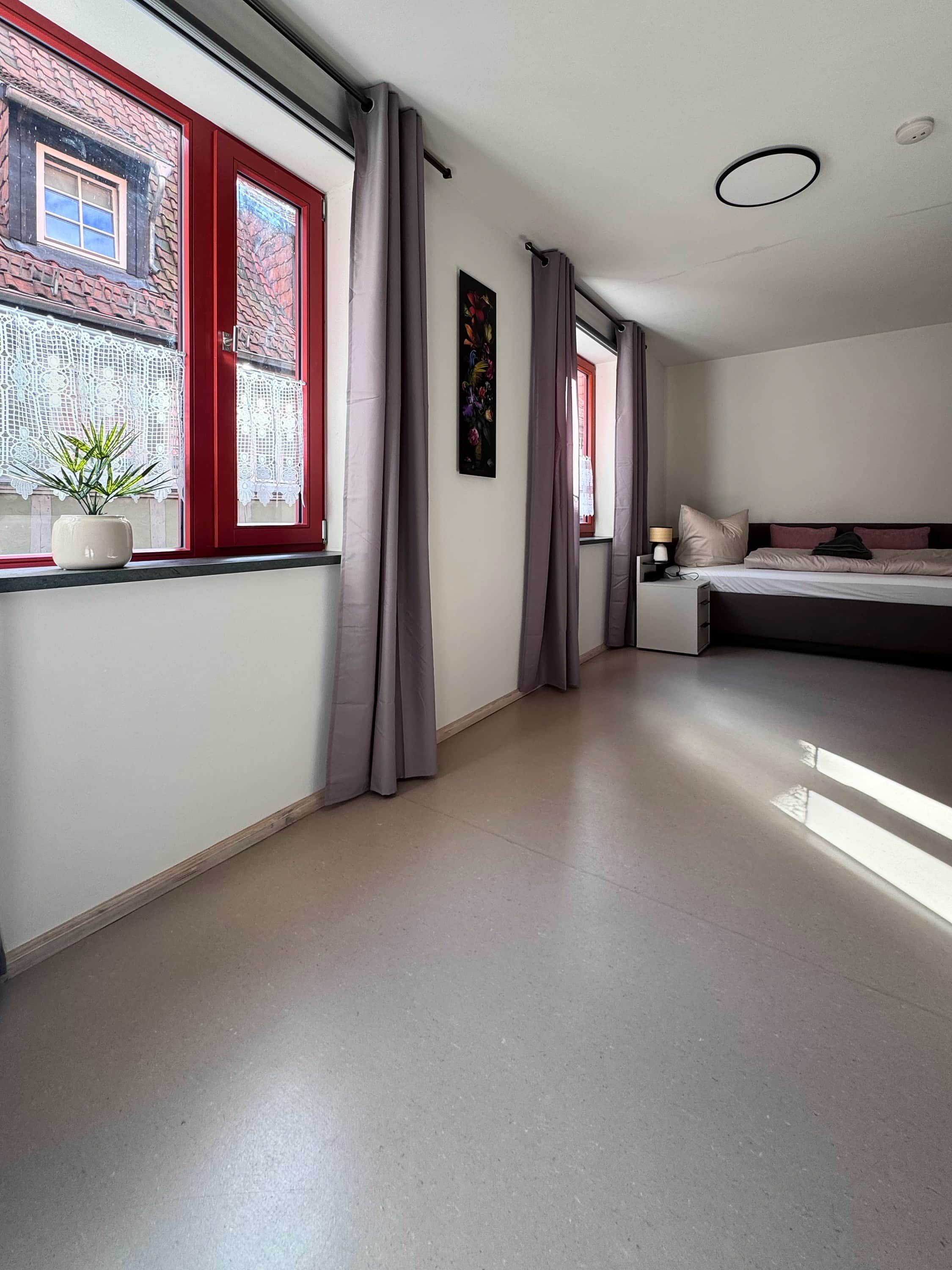 A modern hotel room with a large bed, a window with red shutters, and a potted plant on the windowsill.