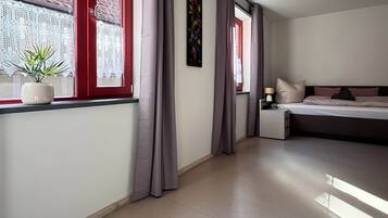 A modern hotel room with a large bed, a window with red shutters, and a potted plant on the windowsill.
