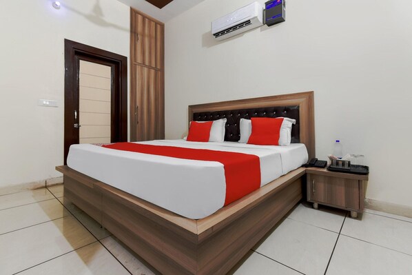 A modern hotel room with a wooden bed frame, a red and white bedspread, a small bedside table with a phone and a bottle, and a wall-mounted air conditioner.