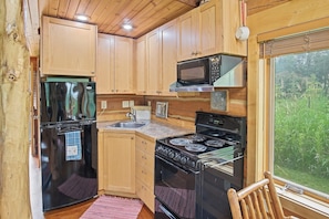 A compact kitchen with wooden cabinets, a refrigerator, a microwave, a stove, and a window with a view of greenery. - Serene 1-bedroom Log Cabin - The St. Croix - on Lake Superior's South Shore (Port Wing)