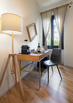 A room with a wooden desk, a chair, a lamp, a framed picture, a bottle of wine, and a window with curtains. - Musart Green Room (Firenze)