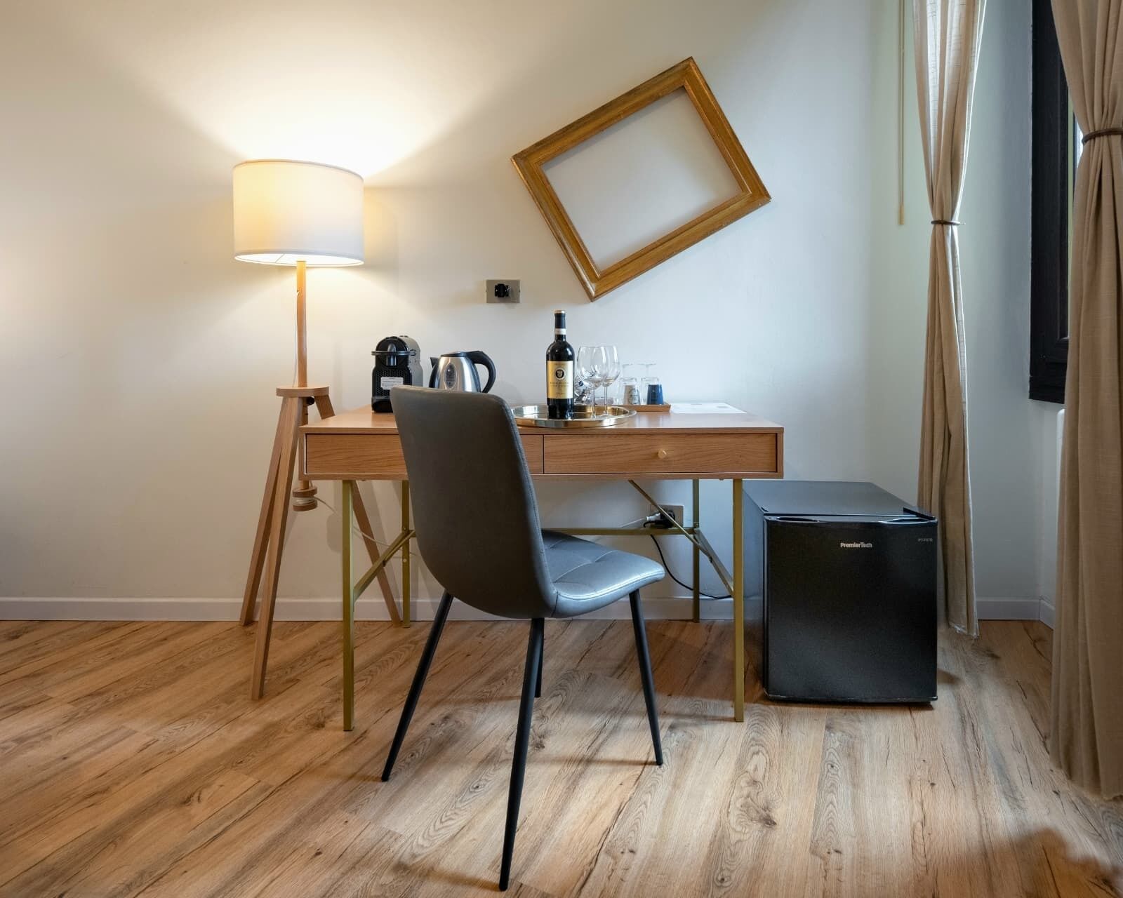 A hotel room with a wooden desk, a chair, a lamp, a refrigerator, and a wall-mounted mirror.