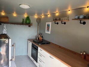 Eine kleine Küche mit Kühlschrank, Backofen und einer Holz-Arbeitsplatte. Lichterketten, eine grüne Vase und ein gerahmtes Bild sind an der Wand zu sehen.