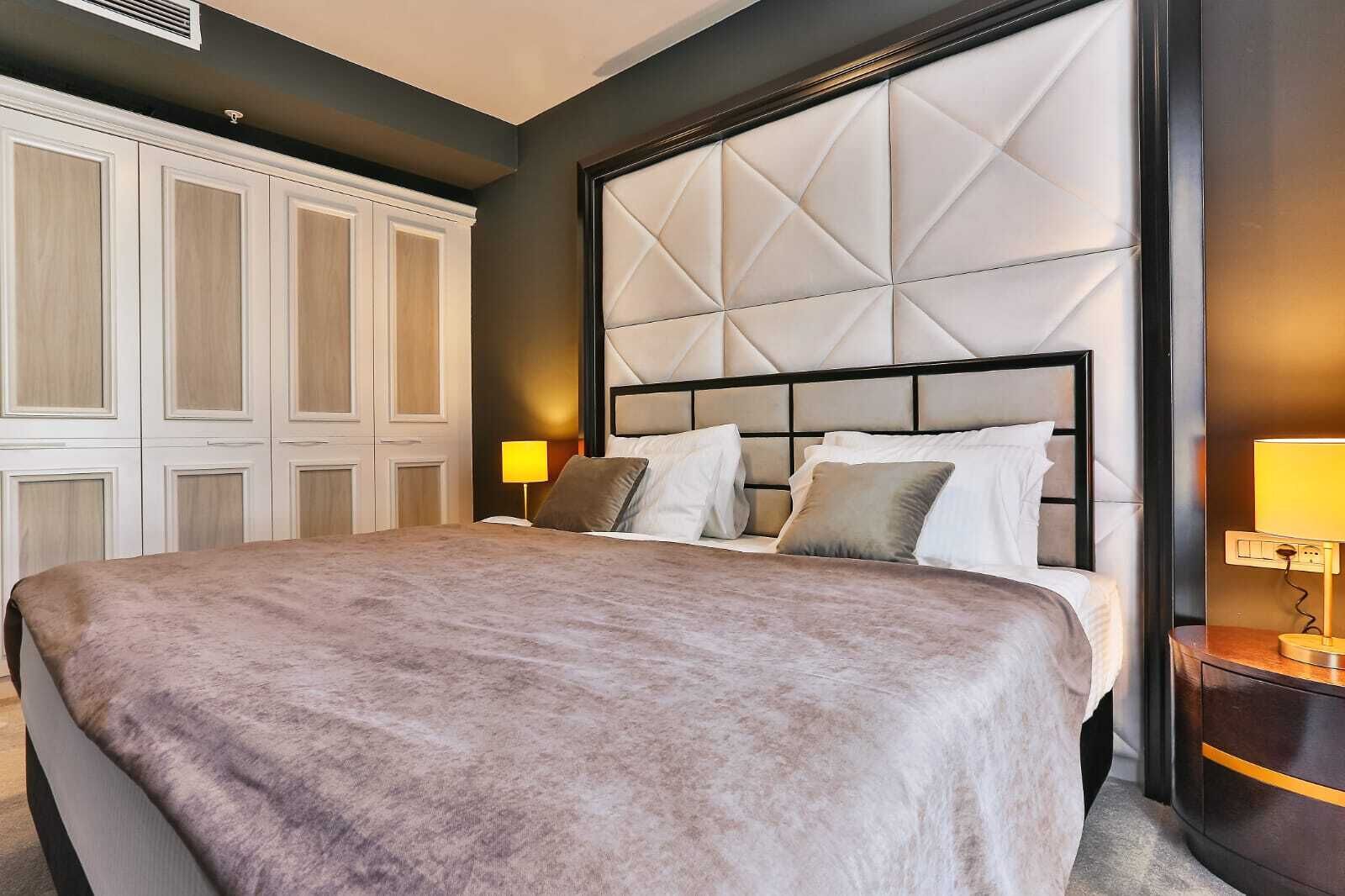 Quarto de hotel com uma cama grande, cabeceira com padrão geométrico, guarda-roupa e mesa de cabeceira com abajur.