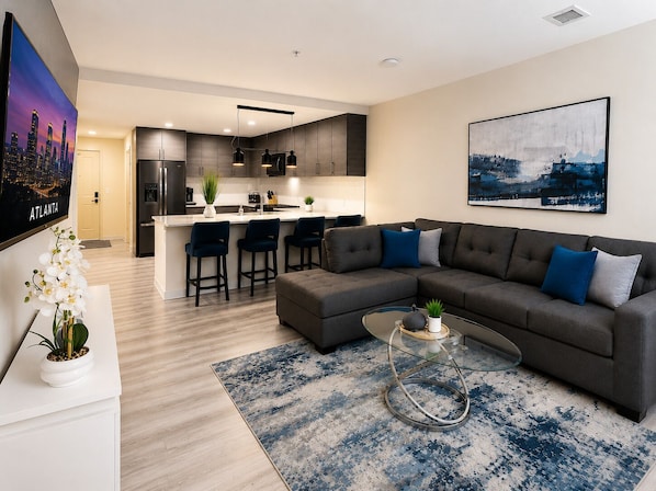 Een moderne woonkamer met een hoekbank, een glazen salontafel en een keuken op de achtergrond.