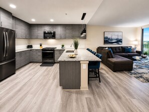 A modern kitchen with a central island, dark cabinetry, and a dining area with a sofa and chairs. - City Skyline Oasis | 2BR Highrise | Year-Round Pool |Balcony | State Farm Arena (Atlanta)