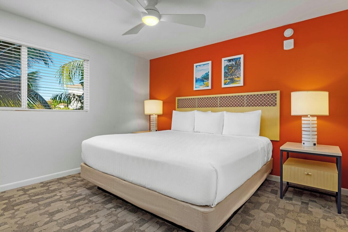 Une chambre d’hôtel avec un lit, deux tables de chevet, un ventilateur de plafond et des œuvres d’art accrochées au mur.