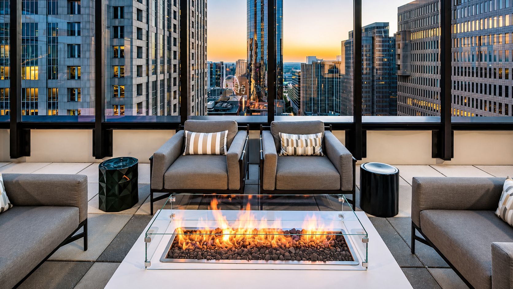 A modern rooftop lounge with a glass fire pit, two sofas with striped pillows, and a city view.