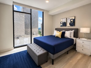 A modern hotel room with a large bed, a nightstand, a lamp, and a view of tall buildings. - Urban City Luxe | Luxury 2BR | Year-Round Pool | Balcony Views| Walk Everywhere (Atlanta)