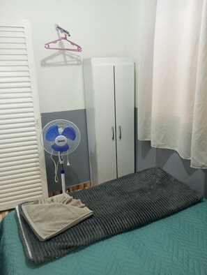 A bedroom with a bed, a standing fan, a wardrobe, and a curtain.