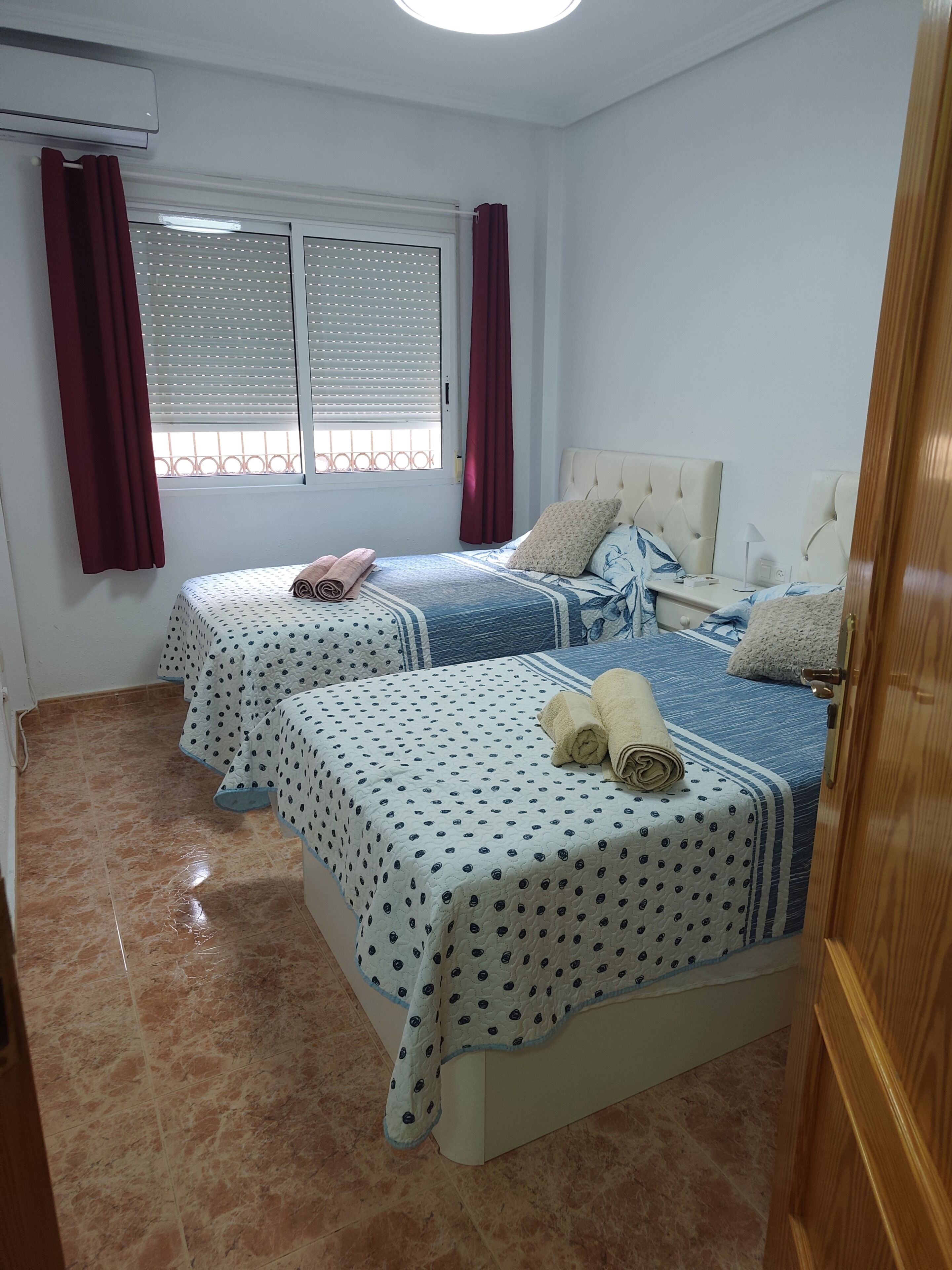 A hotel room with two beds, a window with blinds, a bedside table with a lamp, and a wooden door.