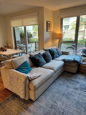 A living room with a beige sectional sofa, a dining table, and chairs. There is a large window providing a view of the outside. - Walk to beach - 2 bedroom apartment - designer furnished - resort style pool (Huntington Beach)