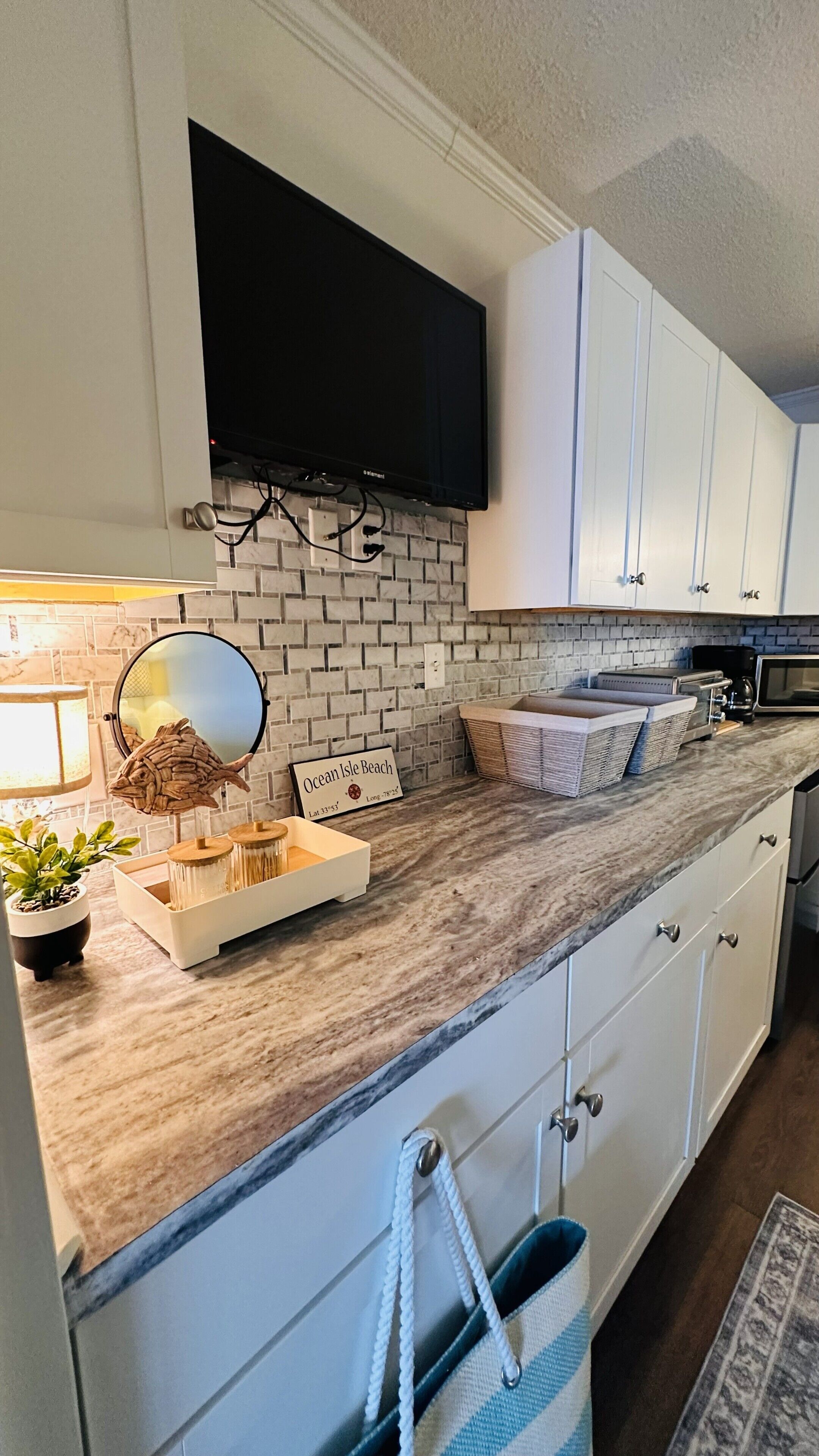 A kitchen with white cabinets, a flat-screen TV mounted above the counter, a brick backsplash, and a sign reading 'Ocean Isle Beach'.