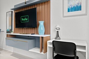 Ein modernes Wohnzimmer mit einem Flachbildfernseher, der das Hulu-Logo zeigt, einem weißen Schreibtisch mit einer Lampe, einem schwarzen Sessel und einer dekorativen Vase.