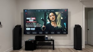 A modern living room with a large flat-screen TV mounted on the wall, a black entertainment unit below, and two black speakers on either side.