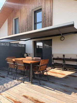 Een houten terras met een eettafel en stoelen, een overdekte patio en een gebouw met een schuin dak.