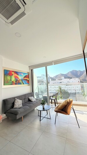 Un salon moderne avec un canapé gris, une table basse ronde et un fauteuil marron. Un tableau coloré orne le mur, et de grandes fenêtres offrent une vue sur des bâtiments et des montagnes.