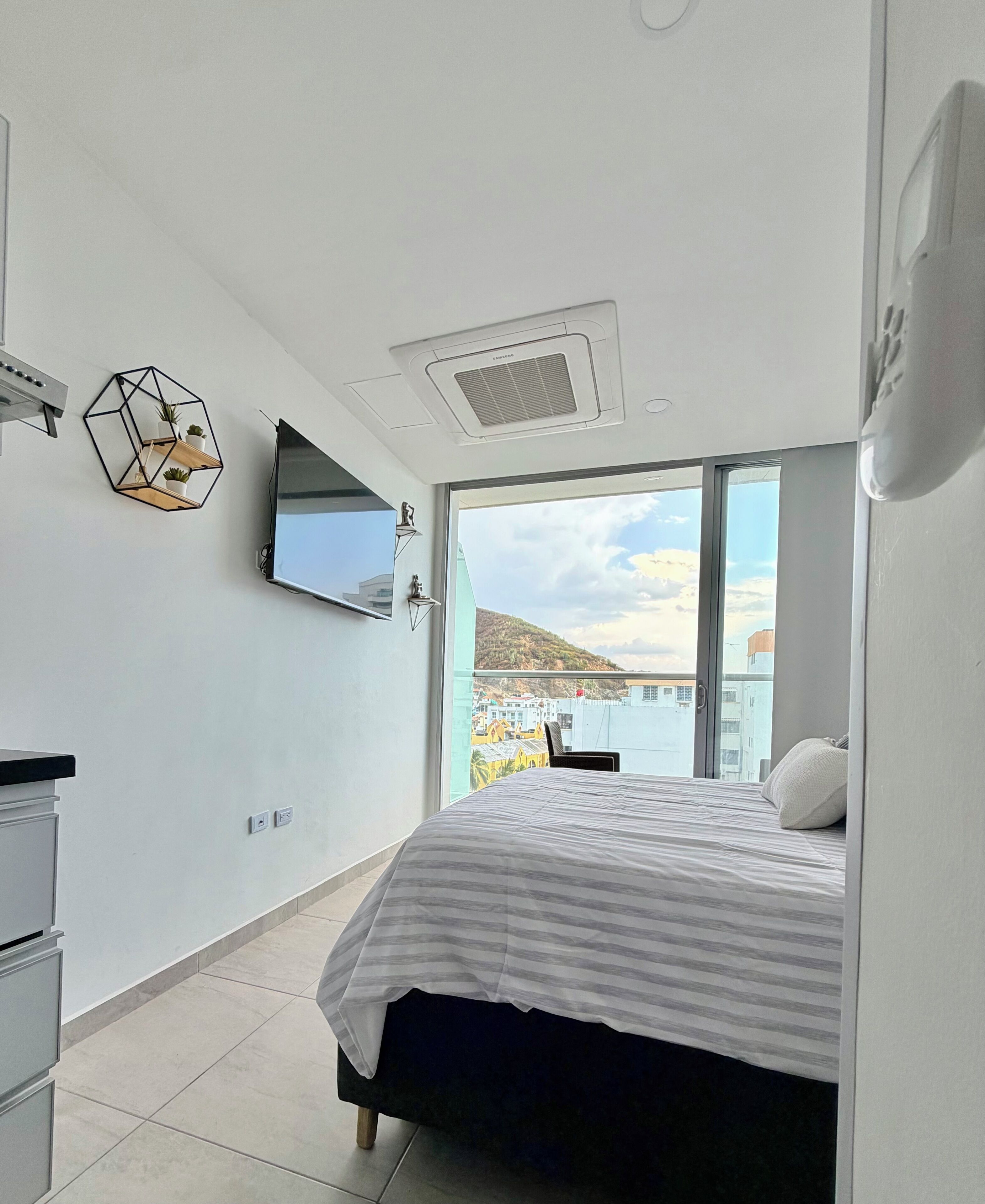 A modern bedroom with a bed, a flat-screen TV, a geometric wall decoration, and a large window offering a view of buildings and hills.