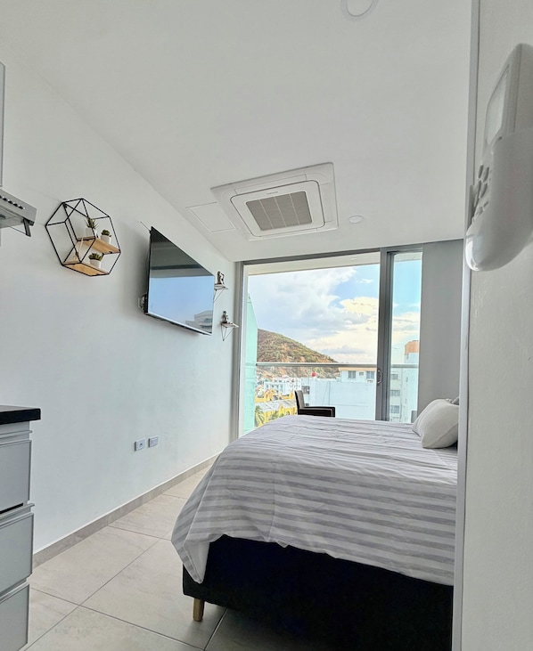 A modern bedroom with a bed, a flat-screen TV, a geometric wall decoration, and a large window offering a view of buildings and hills.