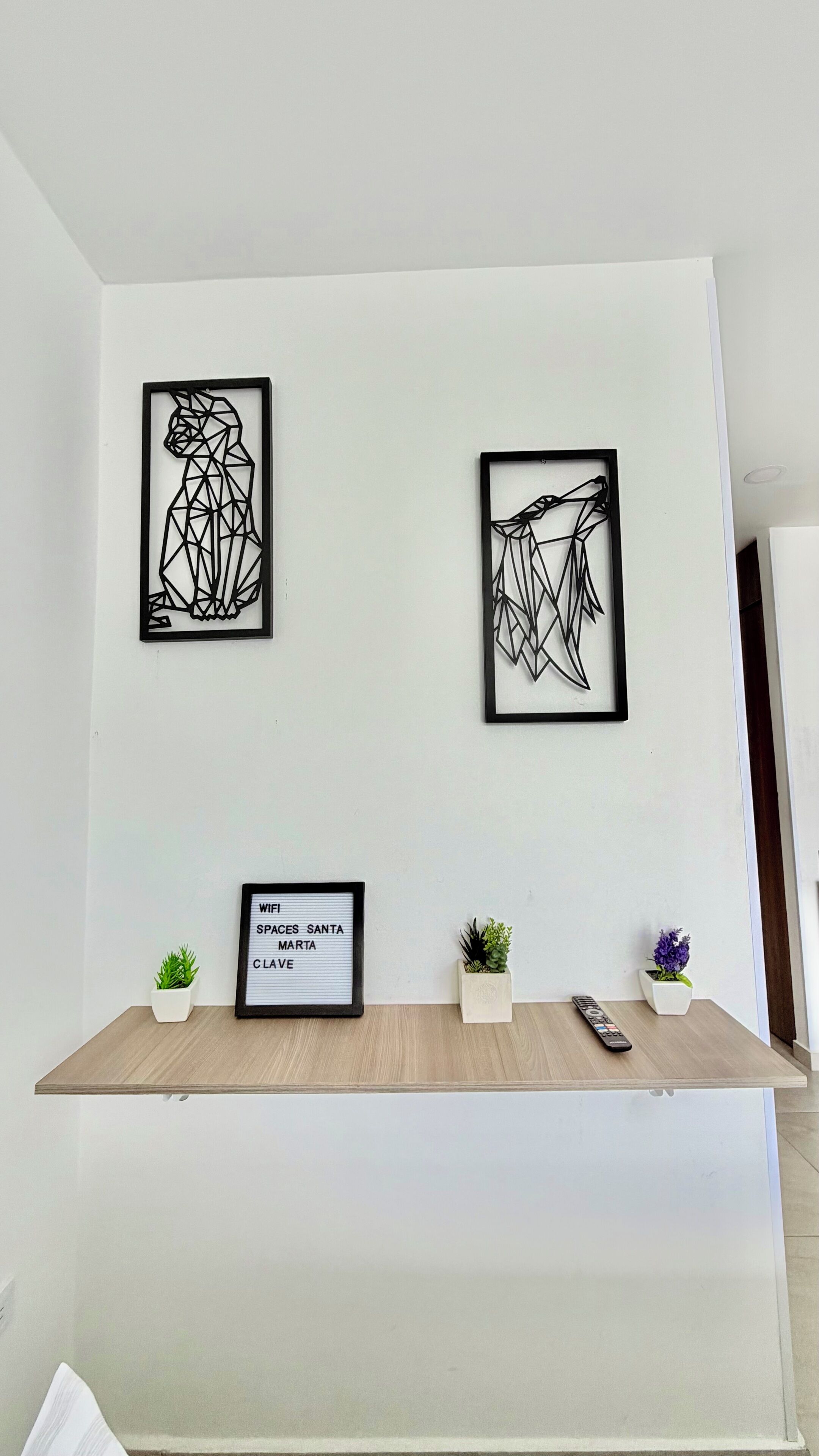 A wall with three framed abstract art pieces and a shelf with a framed sign, two small potted plants, and a remote control.