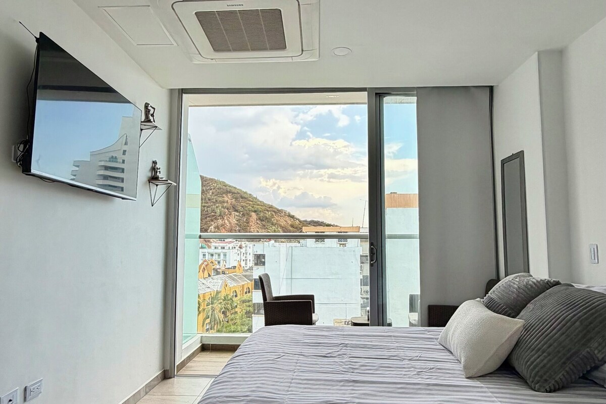 A modern bedroom with a large bed, a flat-screen TV, and a view of a body of water.
