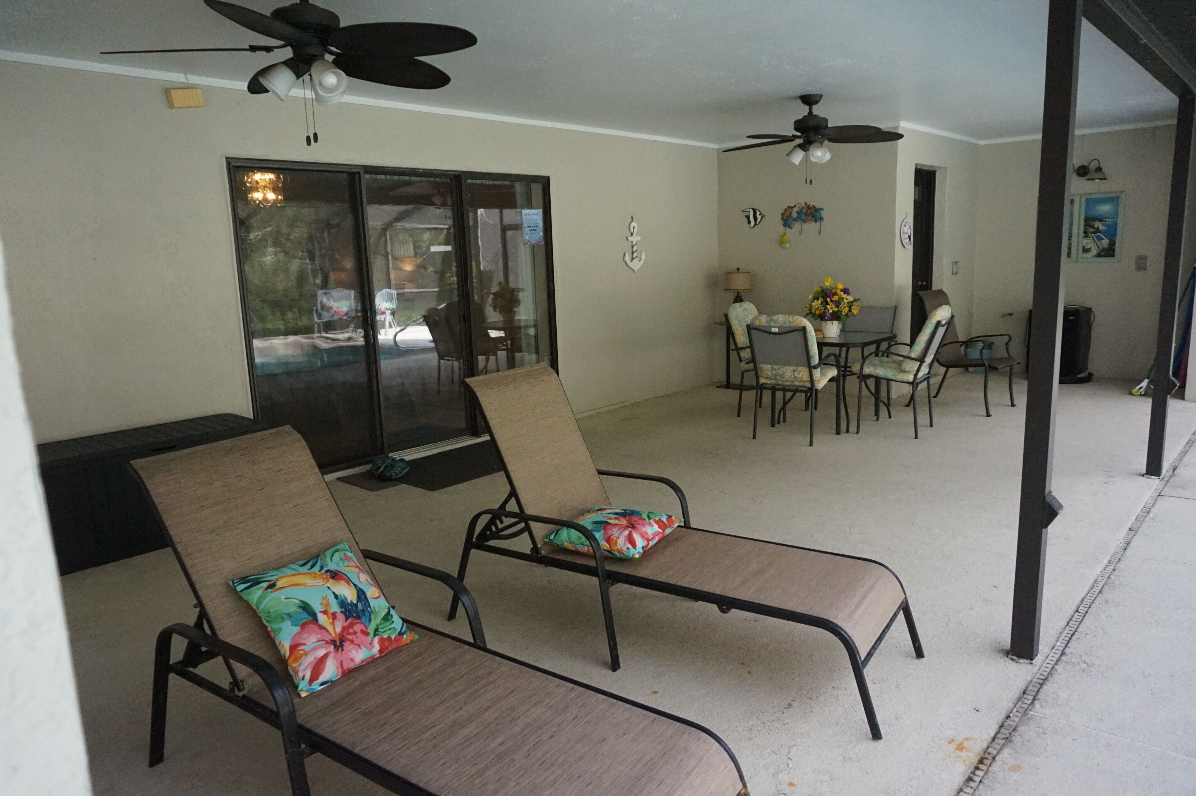 Ein überdachter Patio-Bereich mit zwei Lounge-Sesseln, einem Tisch mit Stühlen, Deckenventilatoren und Blick durch eine Glastür.