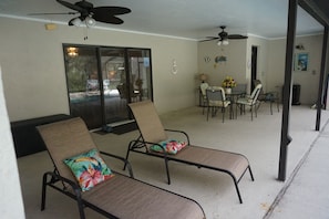 Un patio techado con dos sillones, una mesa y sillas, ventiladores de techo y vistas a través de un ventanal.