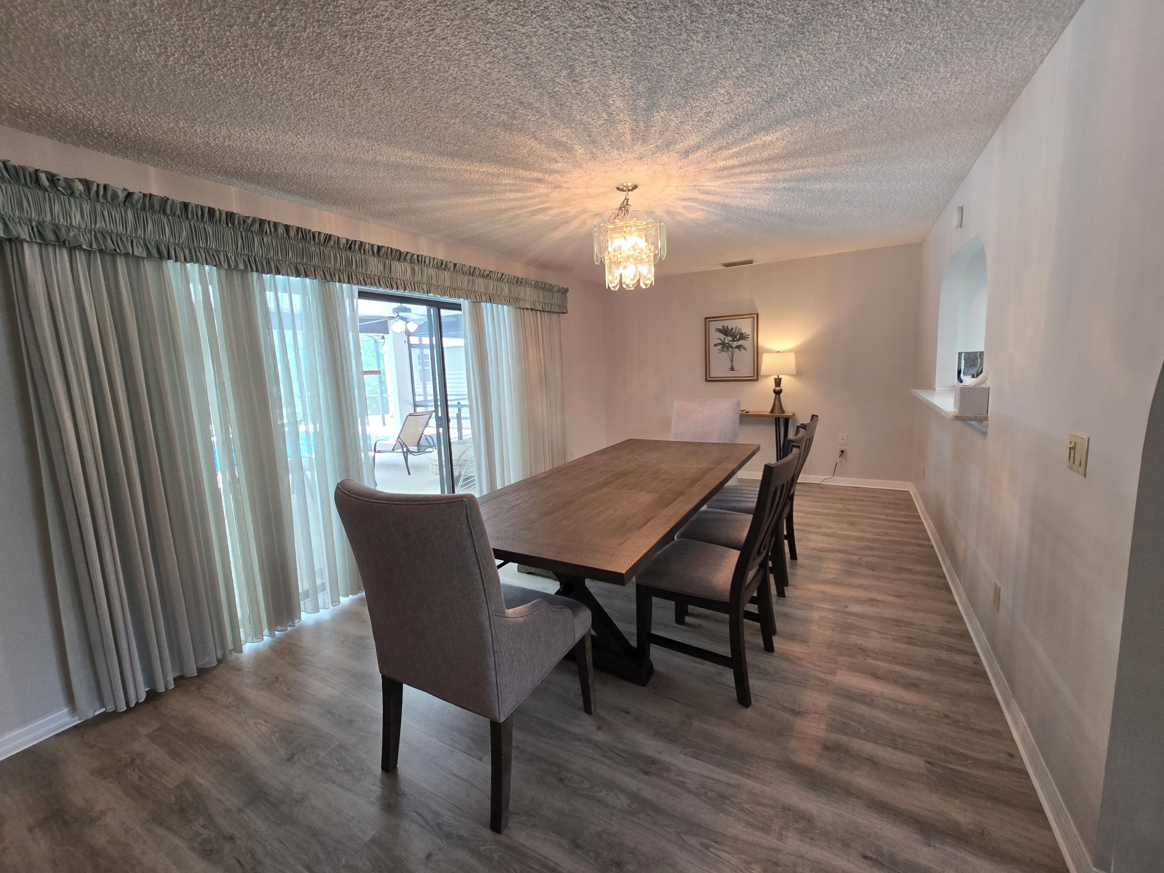 A dining room with a wooden table and chairs, a chandelier, and a view through large windows.