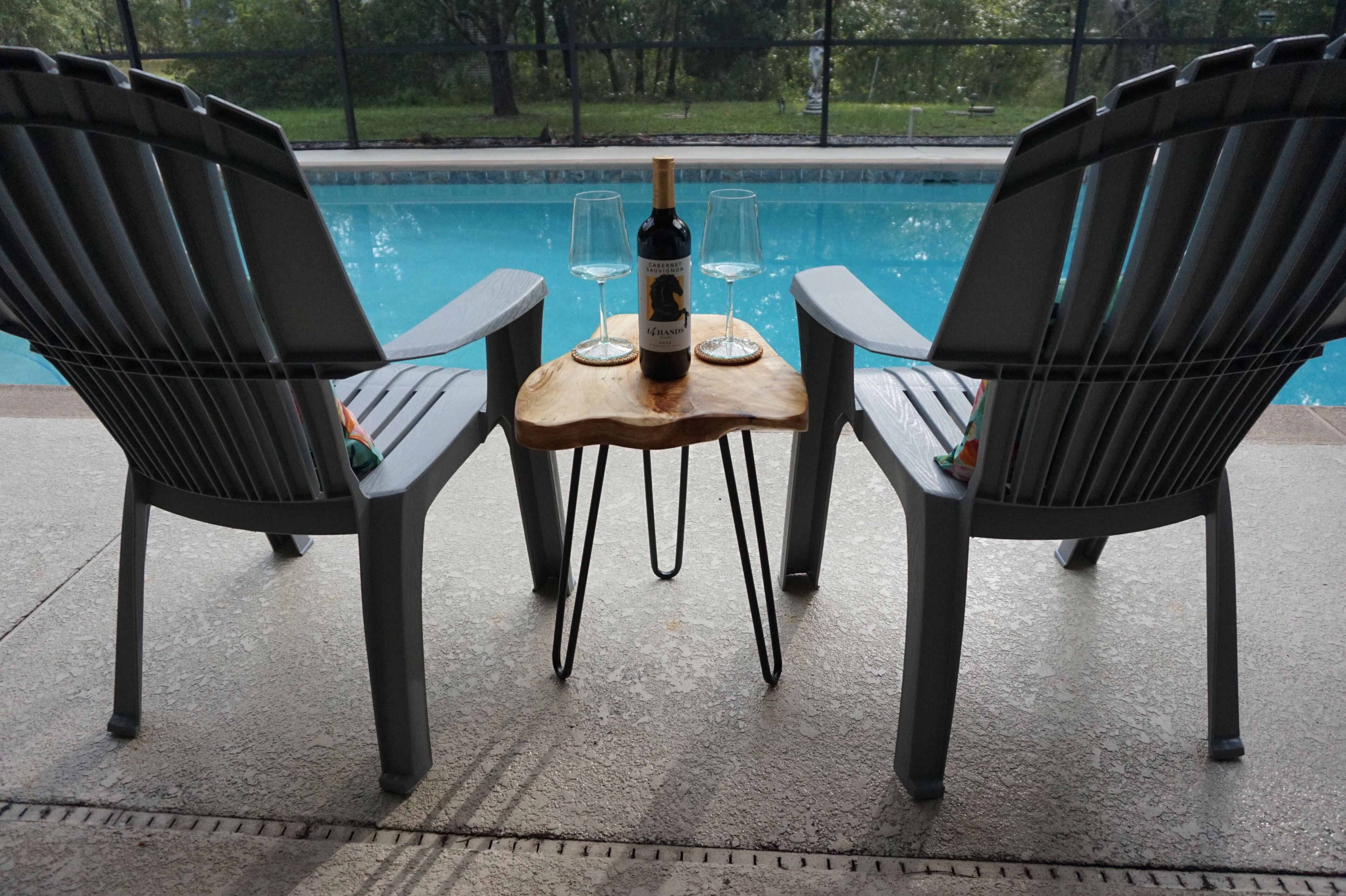 Two Adirondack chairs facing a pool with a wooden table between them, a bottle of wine, and two glasses on the table.