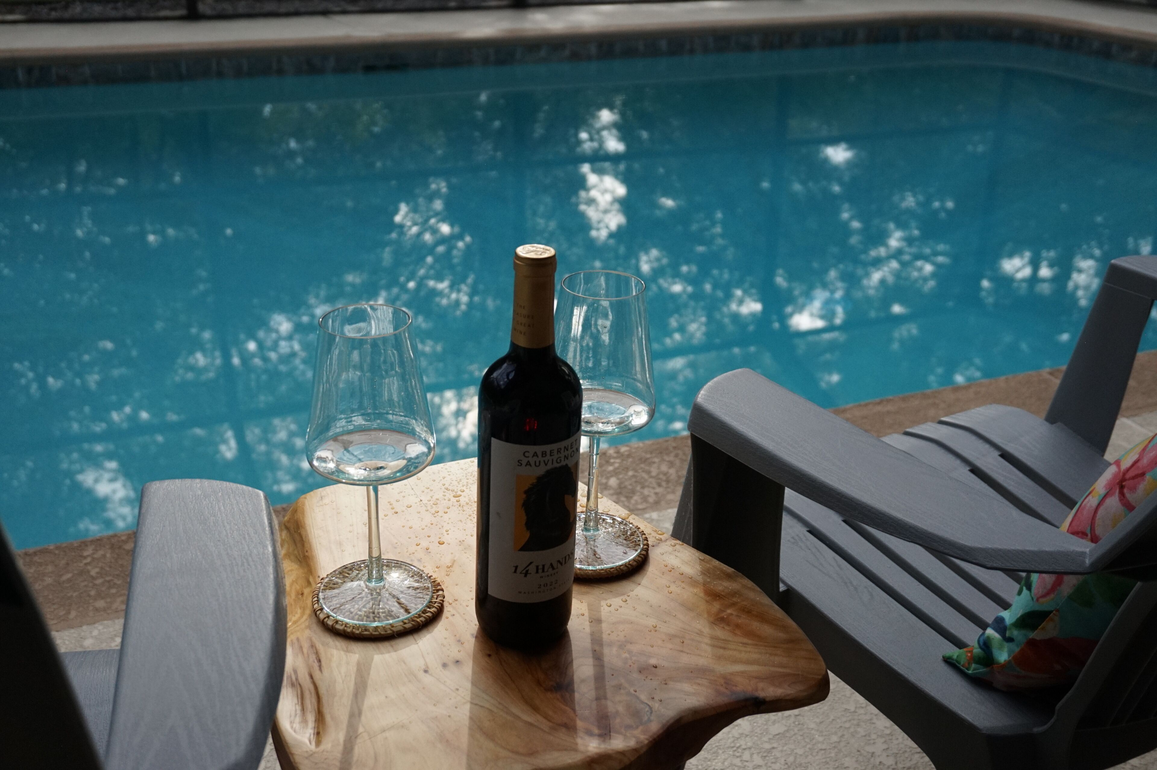 Une scène de bord de piscine avec une bouteille de vin, deux verres et une table en bois.