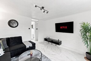 Een moderne woonkamer met een zwarte bank, een glazen salontafel, een wandgemonteerde televisie waarop 'NETFLIX' staat, een zwarte tv-kast, een potplant en een klok aan de muur.
