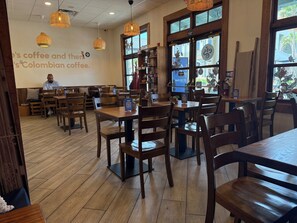 Interior de una cafetería con mesas y sillas de madera, una pared con un anuncio de café y una ventana con vistas al exterior.