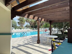 Un área de piscina techada con sillones de descanso y una mesa, rodeada de palmeras.