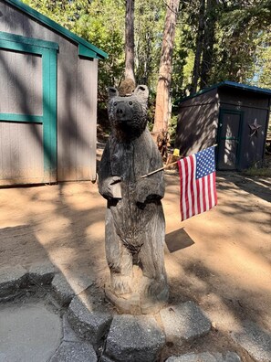 Una escultura de madera de un oso sosteniendo una bandera, rodeado de árboles y un edificio con techo verde.