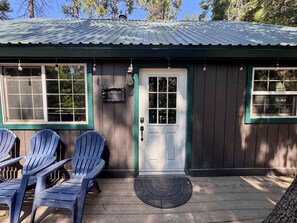 A small wooden building with a metal roof, a white door, and two windows. There are three blue plastic chairs outside. - Foxtail Cabin - Walk to Town & West Village (Shaver Lake)