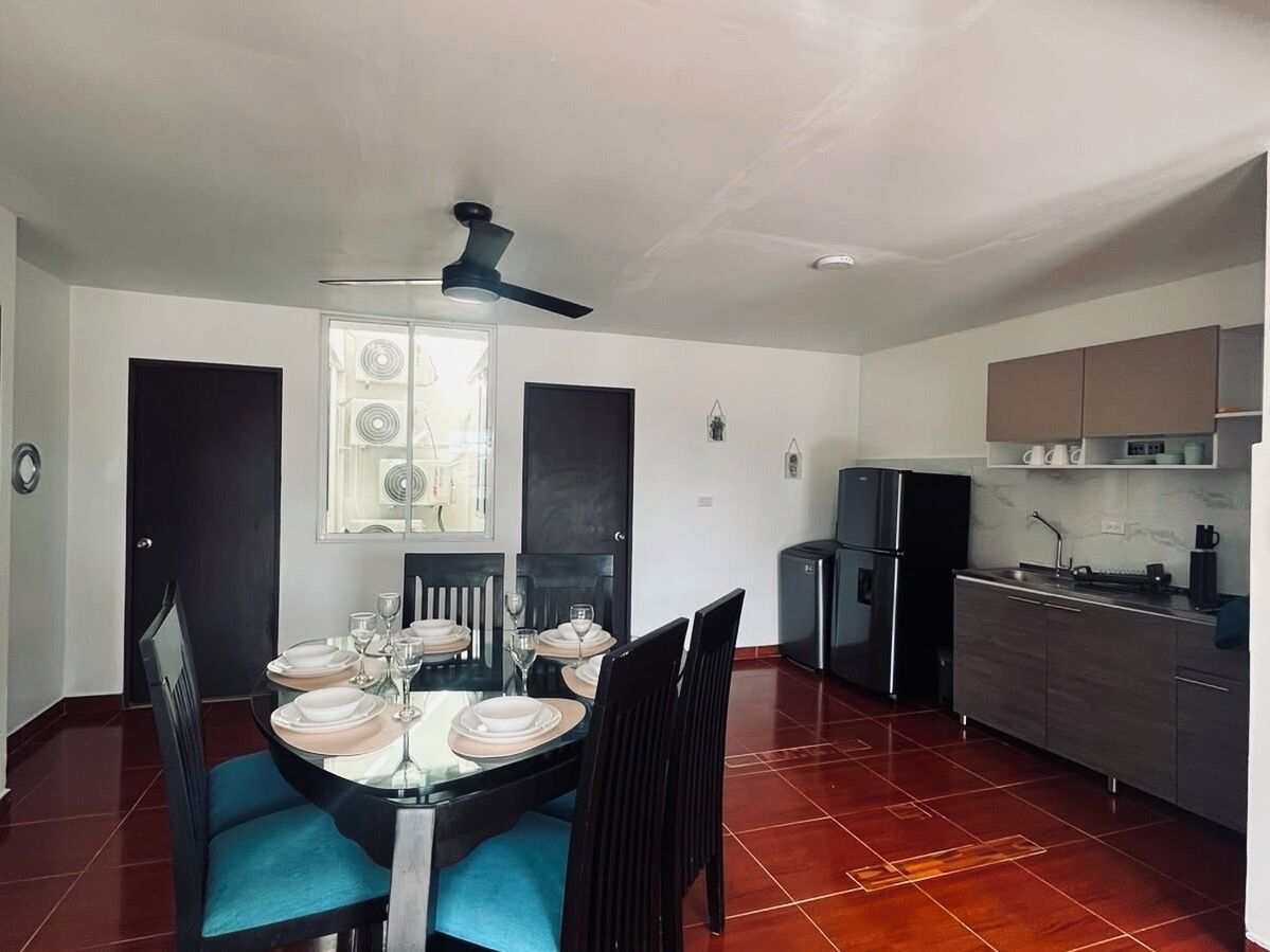 A modern kitchen with a dining area, featuring a round table set for four, a ceiling fan, and a refrigerator.