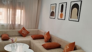 Een woonkamer met een beige hoekbank, een witte salontafel en een witte lamp. Er hangen drie kunstwerken in lijsten aan de muur.
