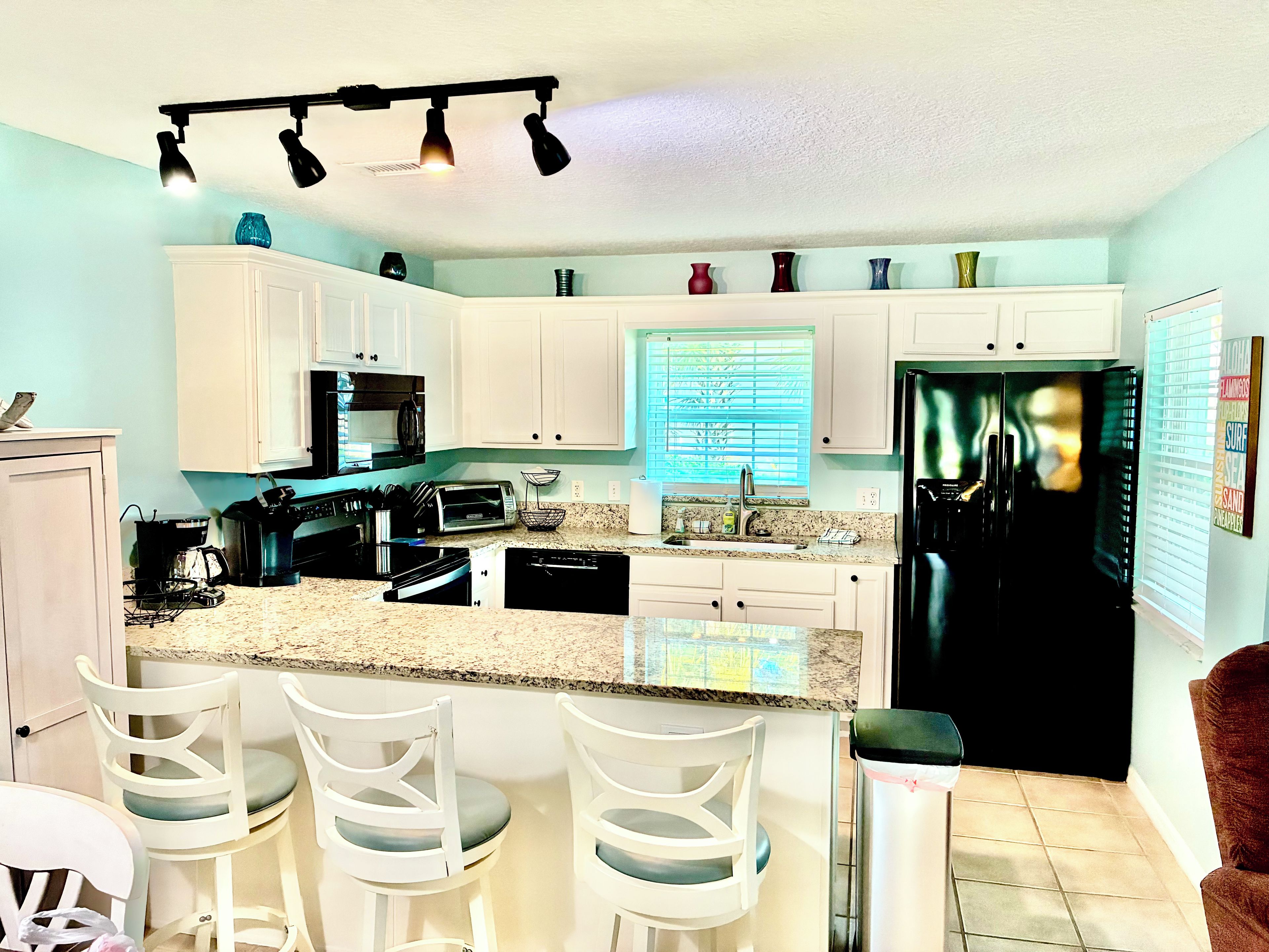 A kitchen with white cabinets, a black refrigerator, a granite countertop, and a window with blinds.
