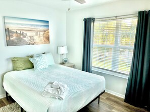 Ein Schlafzimmer mit einem Bett, Kissen, einer Nachttischlampe, einem Fenster mit Vorhängen und einem Bild an der Wand.