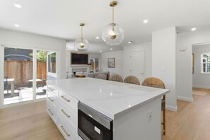 A modern kitchen with a large island, pendant lighting, and a sliding glass door leading to an outdoor area. - Chic 3BR w/ Private Fenced Backyard Oasis (Foster City)