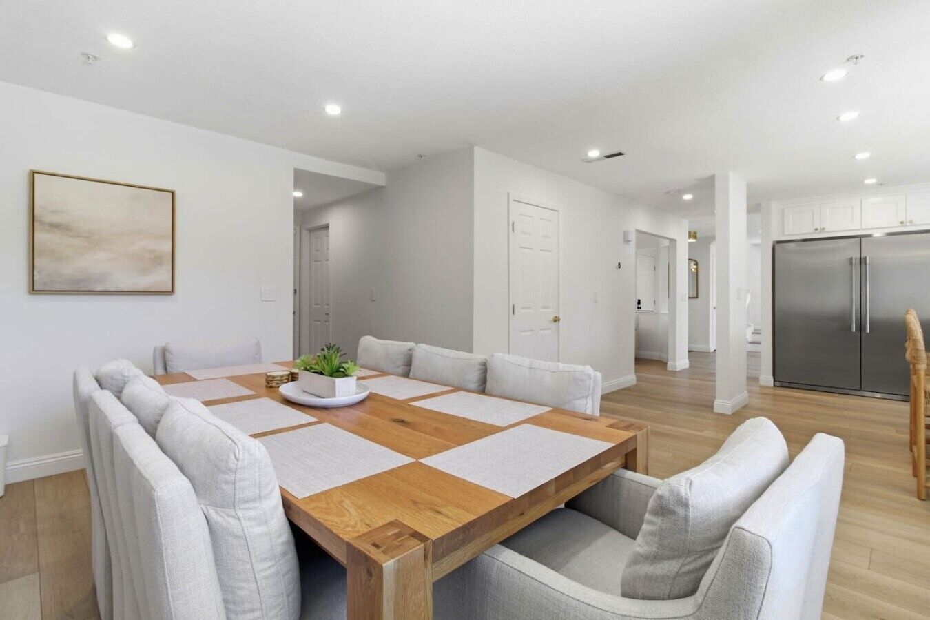 A modern dining area with a wooden table and cushioned chairs, a potted plant, and a minimalist kitchen in the background.