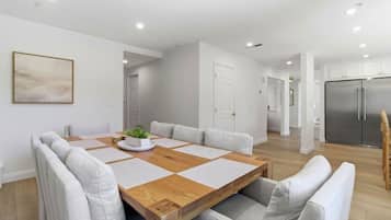 A modern dining area with a wooden table and cushioned chairs, a potted plant, and a minimalist kitchen in the background.