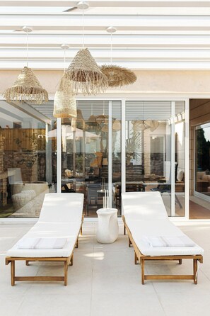 Un área de descanso al aire libre moderna, con dos sillones de madera, una lámpara colgante de fibra y un ventanal que da a un espacio interior.