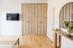 Una habitación con piso de madera, un televisor montado en la pared, una puerta de madera con un panel tejido y una consola con un florero.