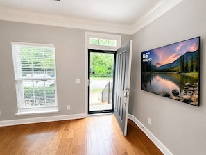 Una habitación con piso de madera, una puerta que da al exterior, una ventana con persianas y un cuadro grande en la pared.