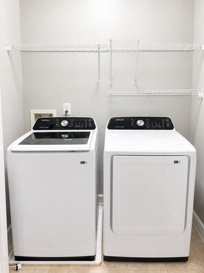 Deux lave-linge à chargement frontal blancs, dotés de panneaux de commande, une étagère blanche au-dessus et une prise électrique blanche sur le mur.