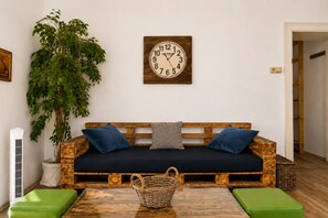 Ein Zimmer mit einem hölzernen Sofa, zwei Kissen, einer Wanduhr, einer Topfpflanze, einem Korbwäschesammler und einer grünen Bank.