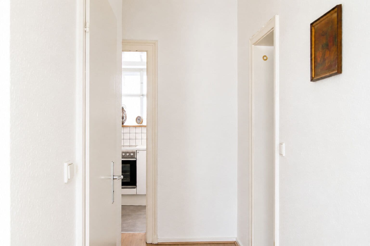 Un pasillo que conduce a una cocina con paredes blancas, una puerta con pomo dorado y un cuadro enmarcado en la pared.